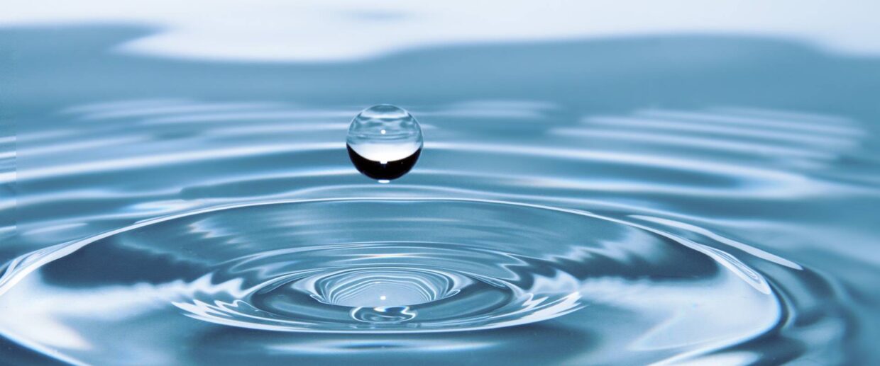 A single drop of water rises from the surface of a blue pool of water.