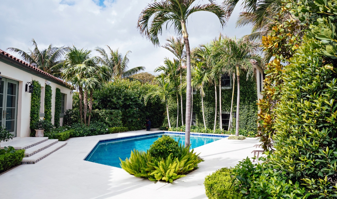 A Palm Beach estate pool area surrounded by mature palms, manicured hedges, ferns, and tropical plantings with a Mediterranean-style home in the background.