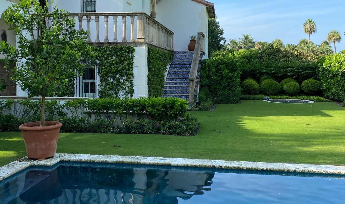 A Palm Beach backyard featuring a blue swimming pool, corner of a house, and meticulously maintained landscaping with pristine turf, trimmed hedges, potted plants, and tropical vegetation.
