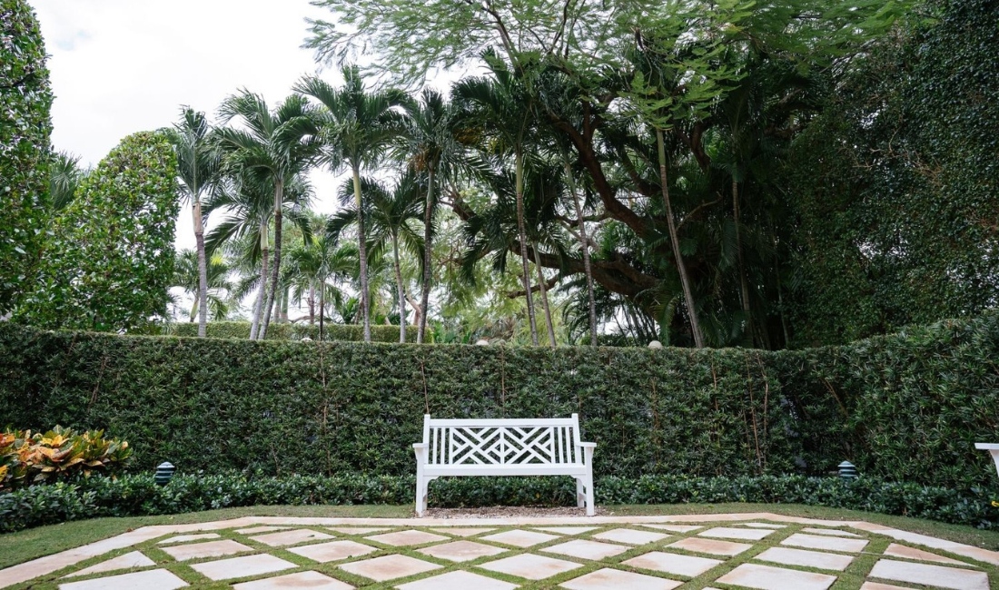 Elegant white garden bench positioned in front of perfectly manicured hedge with geometric stone paver pattern on luxury Palm Beach estate lawn.