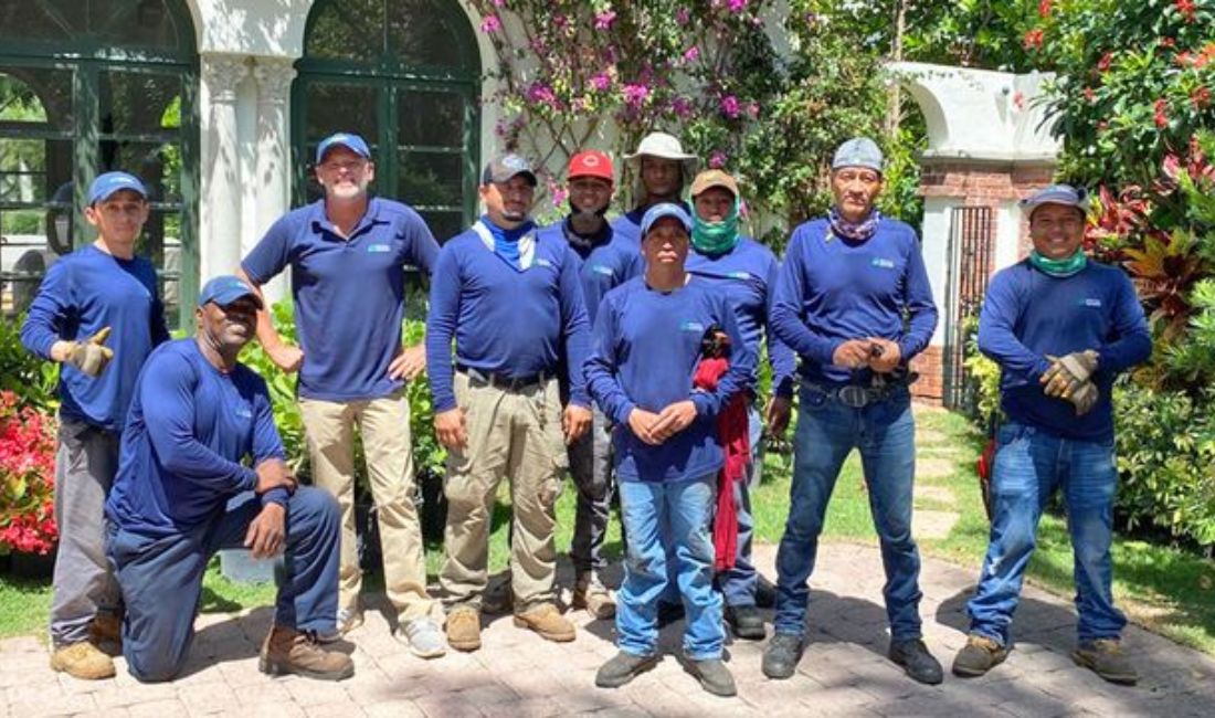 Coastal Gardens professional landscape crew in uniform standing in front of luxury Palm Beach estate with vibrant tropical plants.