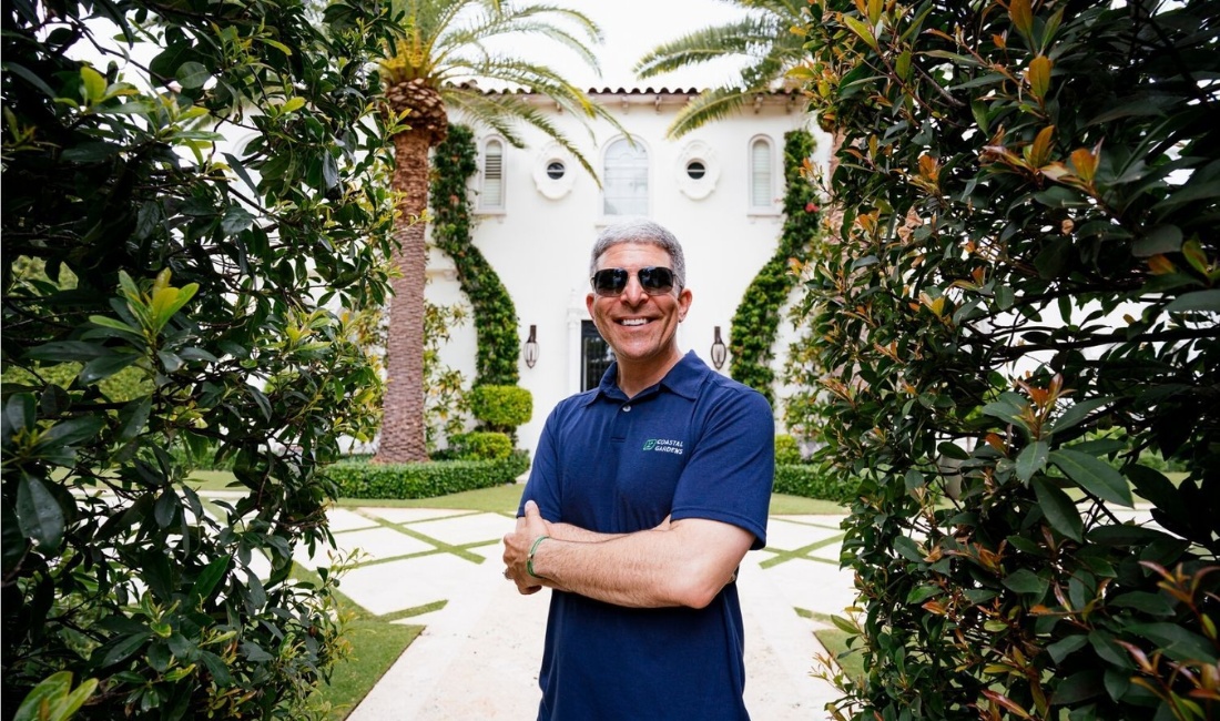 Coastal Gardens professional in navy branded polo standing between mature magnolia hedges on a Palm Beach estate, with date palms and Mediterranean architecture visible in the background.