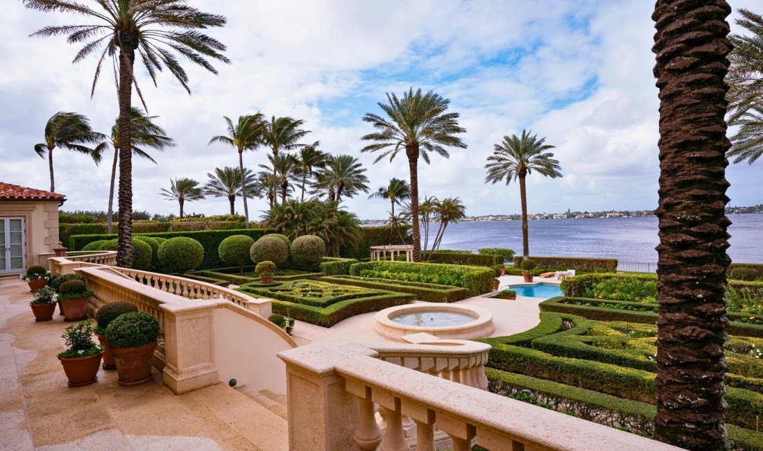 Formal parterre garden on a Palm Beach waterfront estate featuring precision-trimmed boxwood hedges, mature date palms, potted topiaries, stone balustrade, fountain, and Intracoastal views.