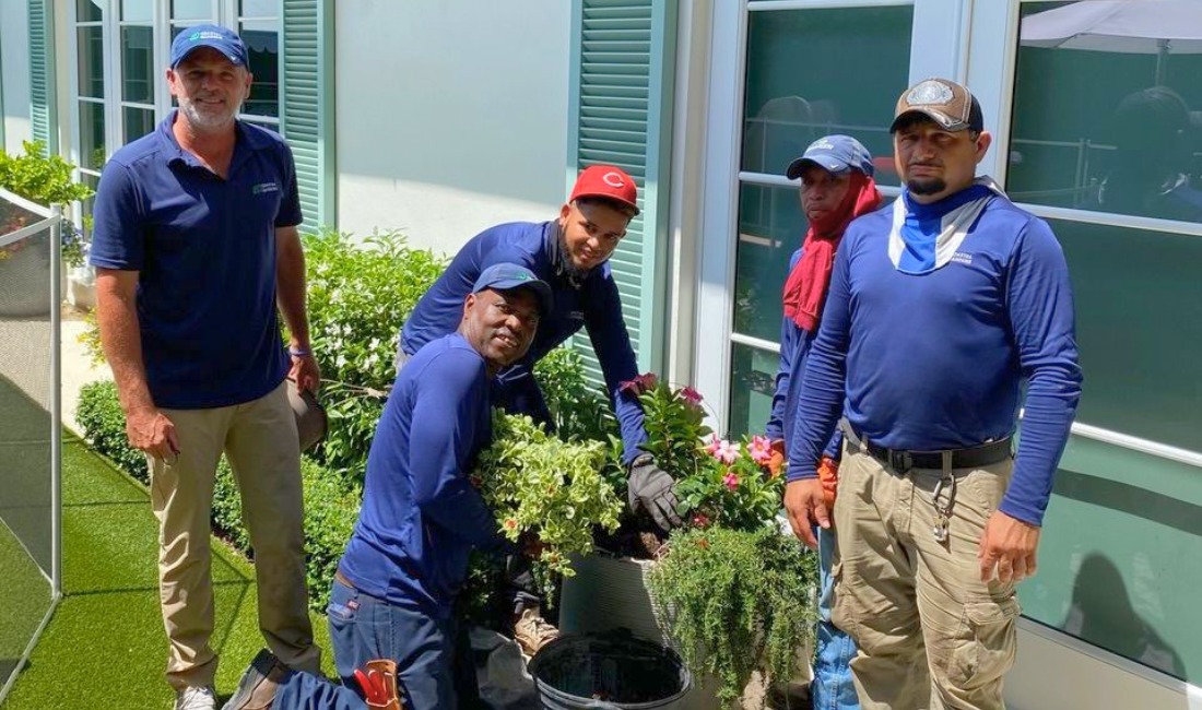 Five Coastal Gardens crew members in blue uniforms posing together while installing a shrub at a Palm Beach estate with a white building in the background.